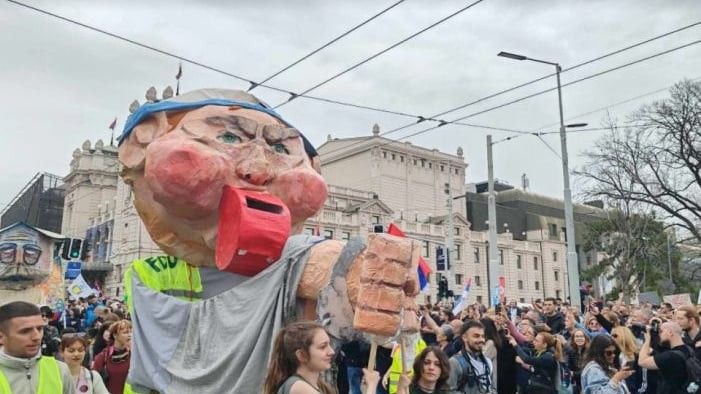 Na čelu kolone bila je lutka u odijelu s kravatom koja na dvije raširene ruke drži ilustraciju krova željezničke stanice u Novom Sadu, dok se u ustima lutke nalazi novac